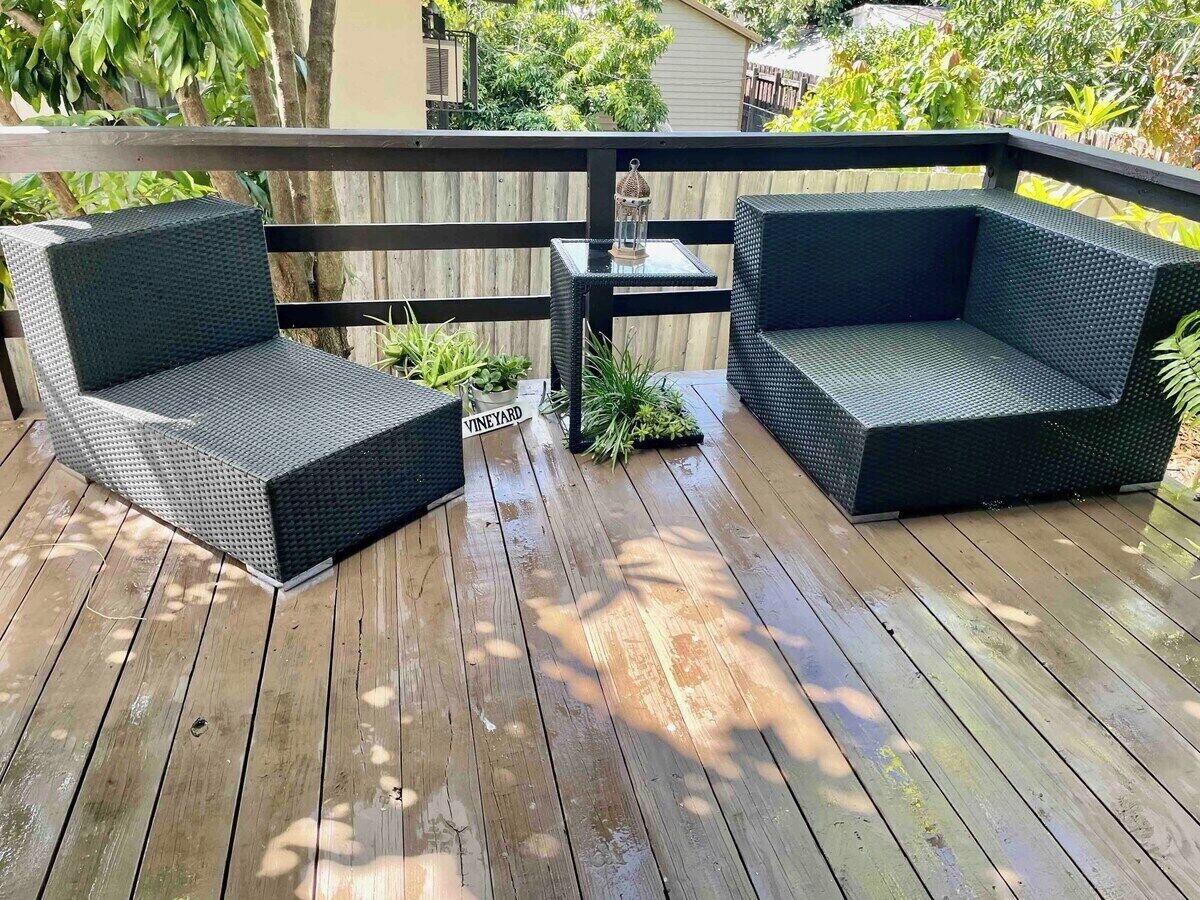 A wooden deck with two dark wicker sofas and a small table, surrounded by a wooden fence and greenery.