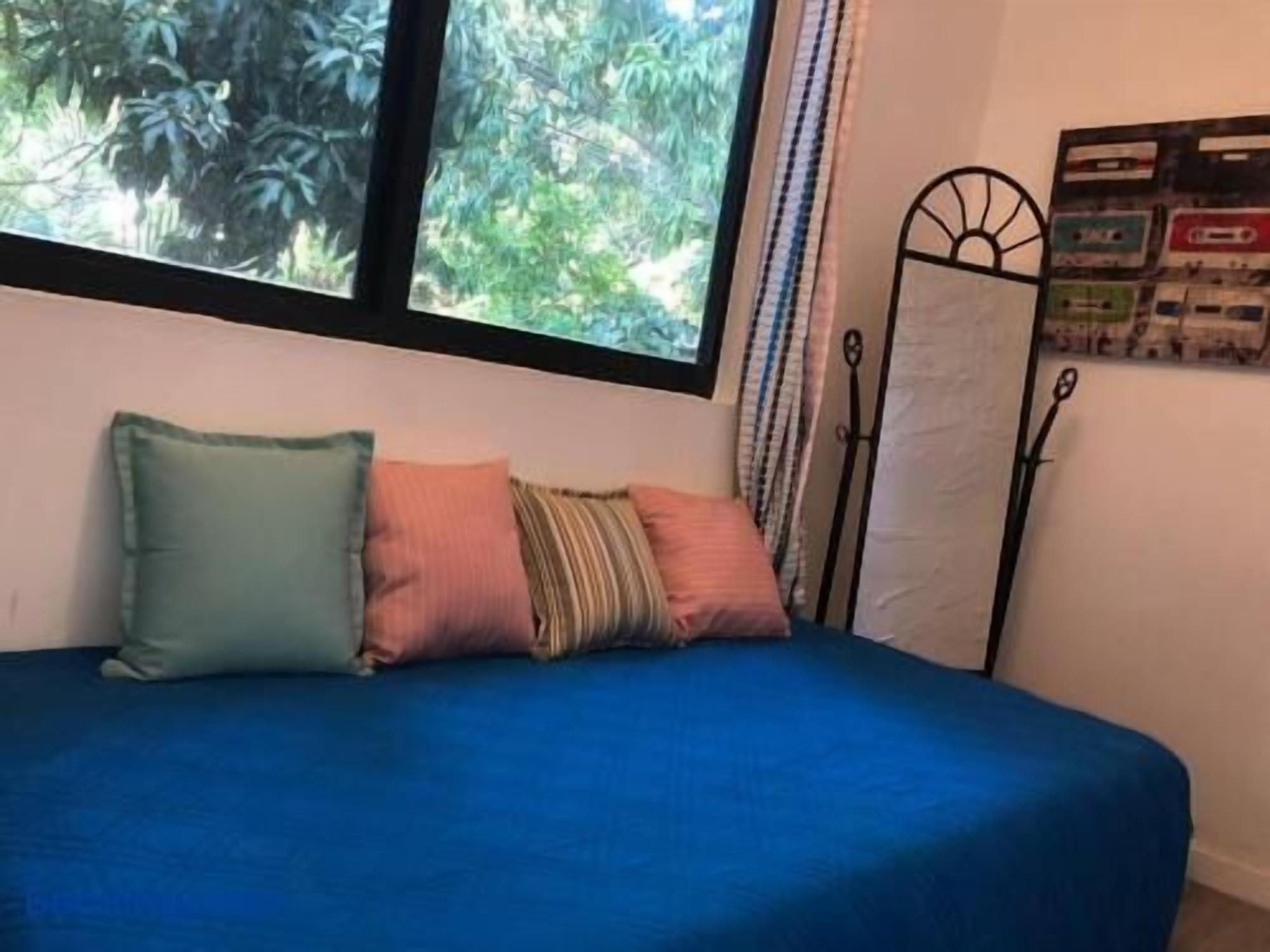 A bed with a blue bedspread and multiple pillows, a window with a view of greenery, and a metal stand with a poster on the wall.