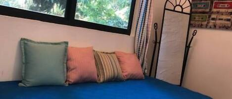 A bed with a blue bedspread and multiple pillows, a window with a view of greenery, and a metal stand with a poster on the wall.