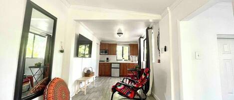 A hallway leading to a kitchen and dining area with a mirror, a patterned cushion, and a rocking chair.