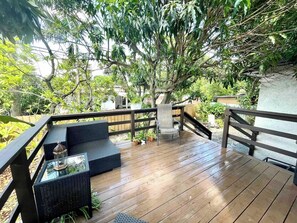 Una terraza de madera con mobiliario exterior, barandilla y un árbol que da sombra.