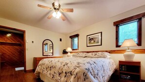 Un dormitorio con piso de madera, una cama con ropa floral, un ventilador de techo, una mesita de noche con una lámpara, una butaca con libros y una ventana con persianas.