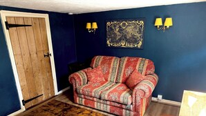 A room with a wooden door, a patterned sofa, a small table, and wall-mounted lamps. - Cosy Victorian Cottage in Crowborough with Log Burner and parking (Crowborough)