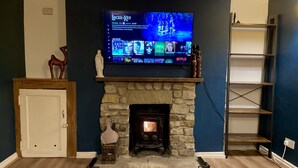 A living room with a stone fireplace, a television mounted above it displaying a Netflix interface, and a wooden cabinet to the left. - Cosy Victorian Cottage in Crowborough with Log Burner and parking (Crowborough)