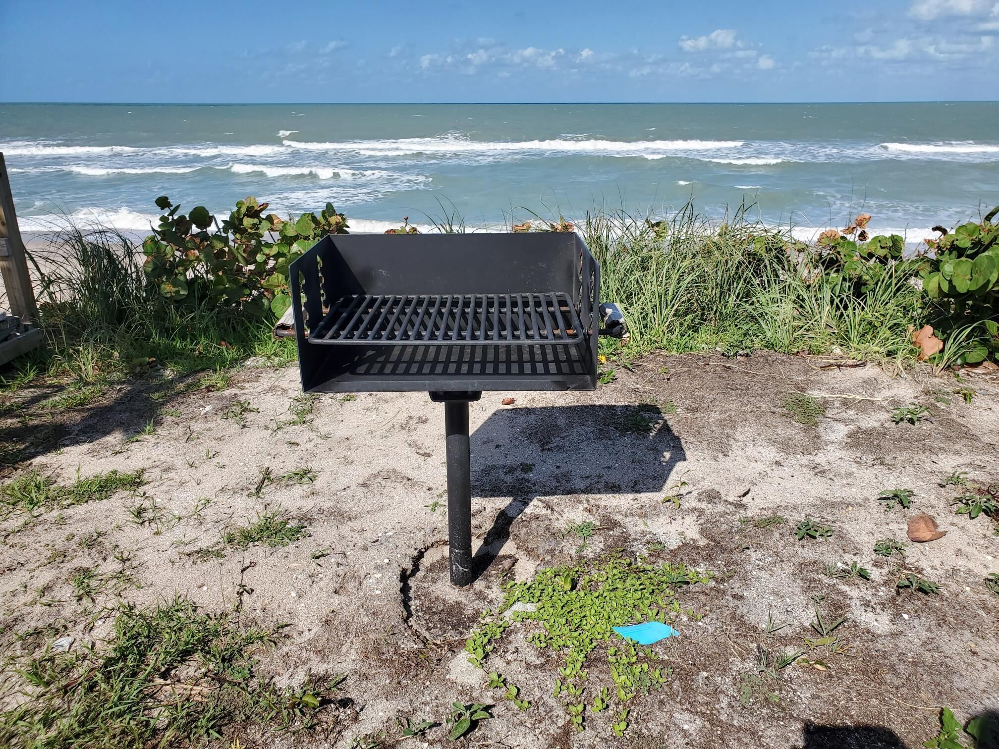 Ein Grill auf sandigem Strand, im Hintergrund das Meer mit Wellen.
