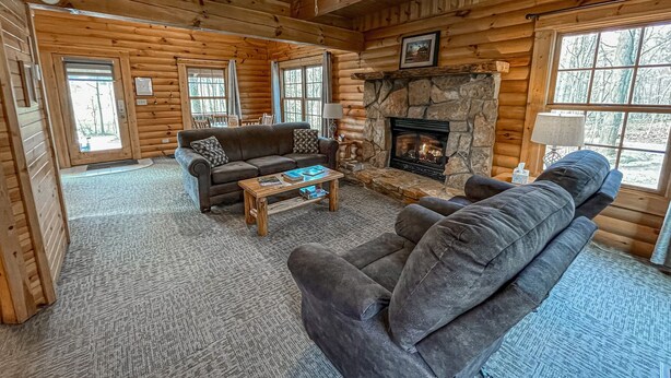 A log cabin interior with a stone fireplace, wooden furniture, and large windows.