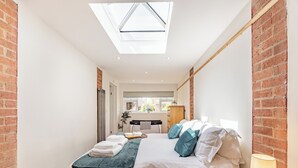Un dormitorio moderno con tragaluces, una cama con almohadas, una mesita de noche con lámpara y vistas a un baño y una cocina al fondo.