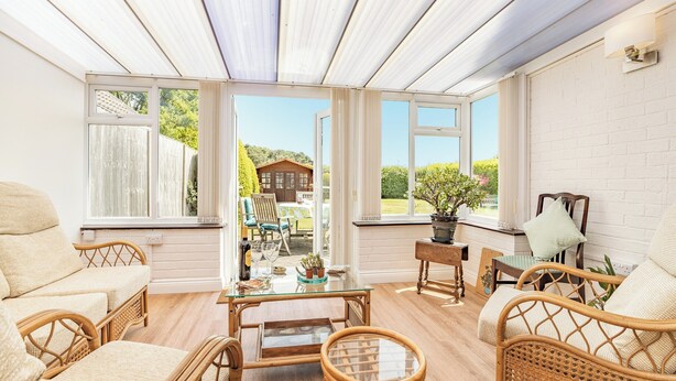 Un solárium con mobiliario de mimbre, ventanas amplias y vistas a una casa y jardín.