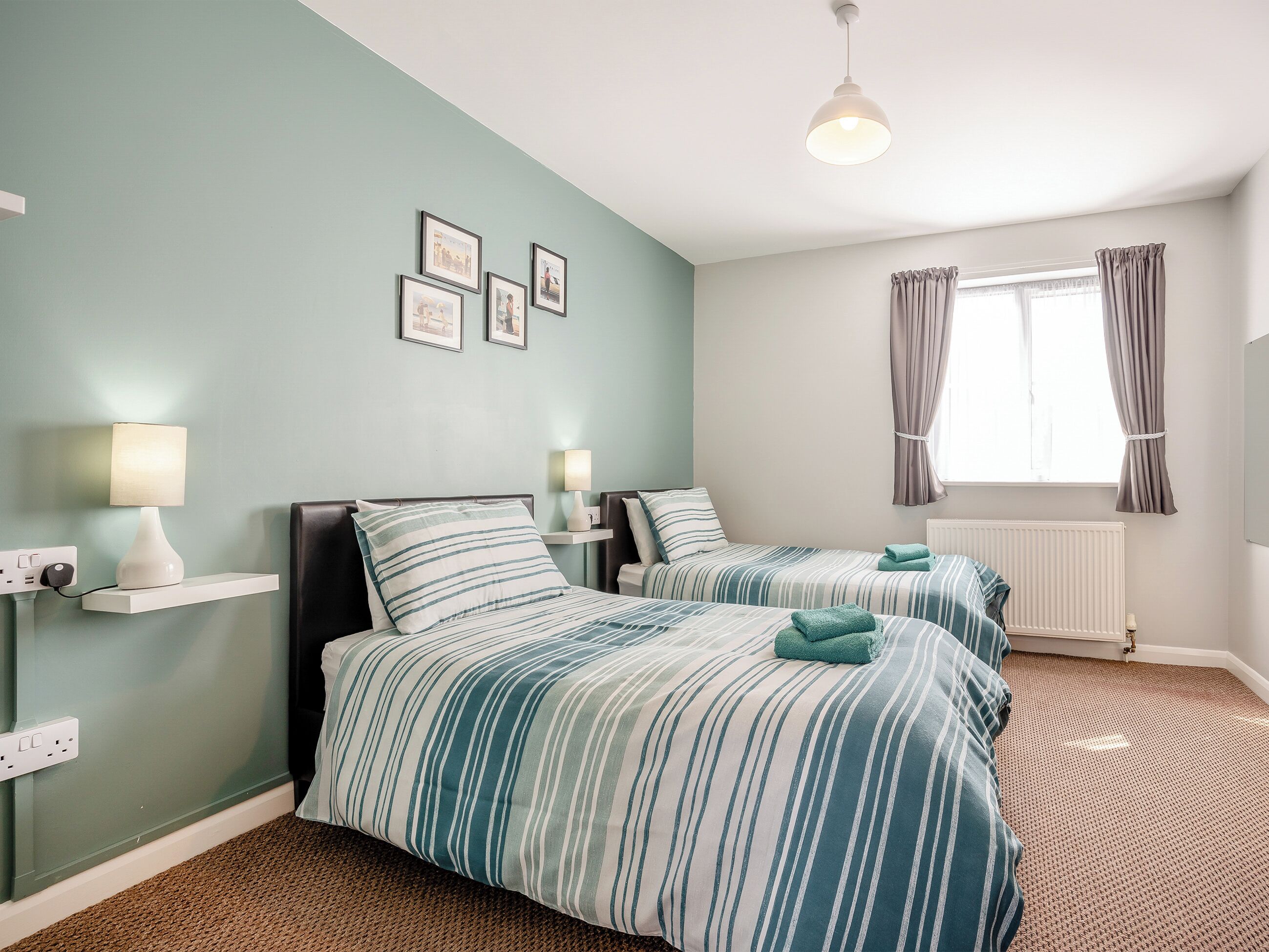 A bedroom with two beds, a window with curtains, a radiator, and wall-mounted lamps.
