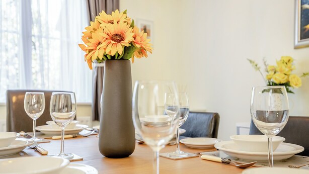 Une table à manger dressée avec de l’argenterie blanche, des verres et des couverts, et un vase rempli de fleurs jaunes placé au centre.