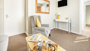 Une chambre d’hôtel moderne avec un fauteuil gris, une table d’appoint en bois sur laquelle se trouve un journal, deux tasses et une cafetière. Un téléviseur à écran plat est fixé au mur, et une petite table avec un vase de fleurs est visible.