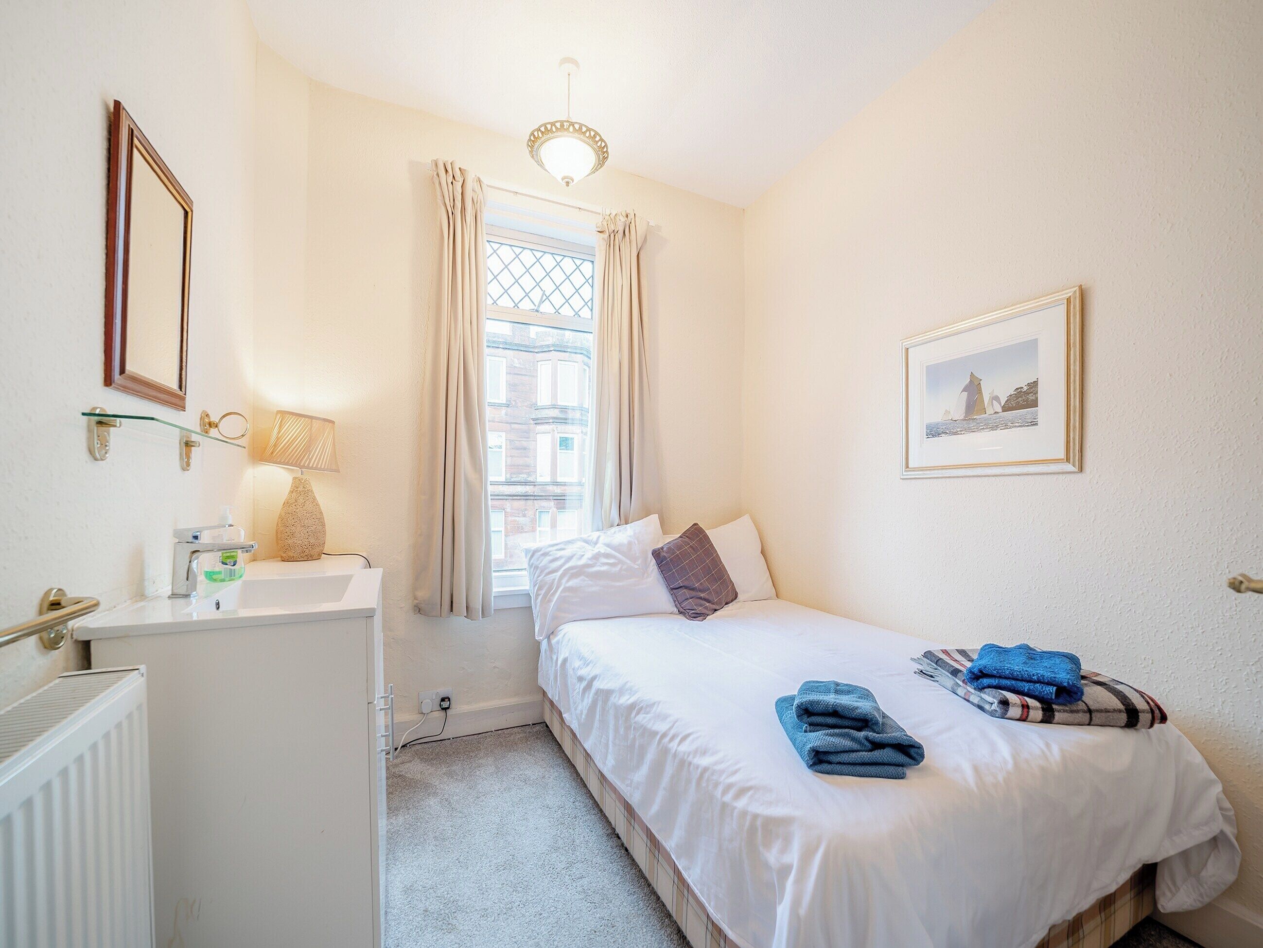 Une chambre d’hôtel avec un lit, une table de chevet avec une lampe, une fenêtre avec des rideaux, un radiateur et un tableau encadré au mur.
