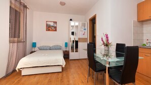 Een kleine kamer met een bed, een glazen eettafel met stoelen, een kitchenette en een deur naar een andere kamer.