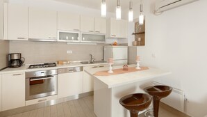 Une cuisine moderne avec des meubles blancs, des appareils en acier inoxydable, un îlot central doté de tabourets, un éclairage suspendu et un appareil de climatisation.