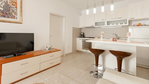 Une cuisine moderne avec des armoires blanches, un four encastré et un four à micro-ondes. On y trouve également un grand téléviseur à écran plat posé sur un meuble en bois, ainsi que deux tabourets de bar au design original.