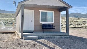 A small, single-story building with a white door and a window, a bench outside, and a clear sky in the background. - Home Base Resort  #5 (Joseph)