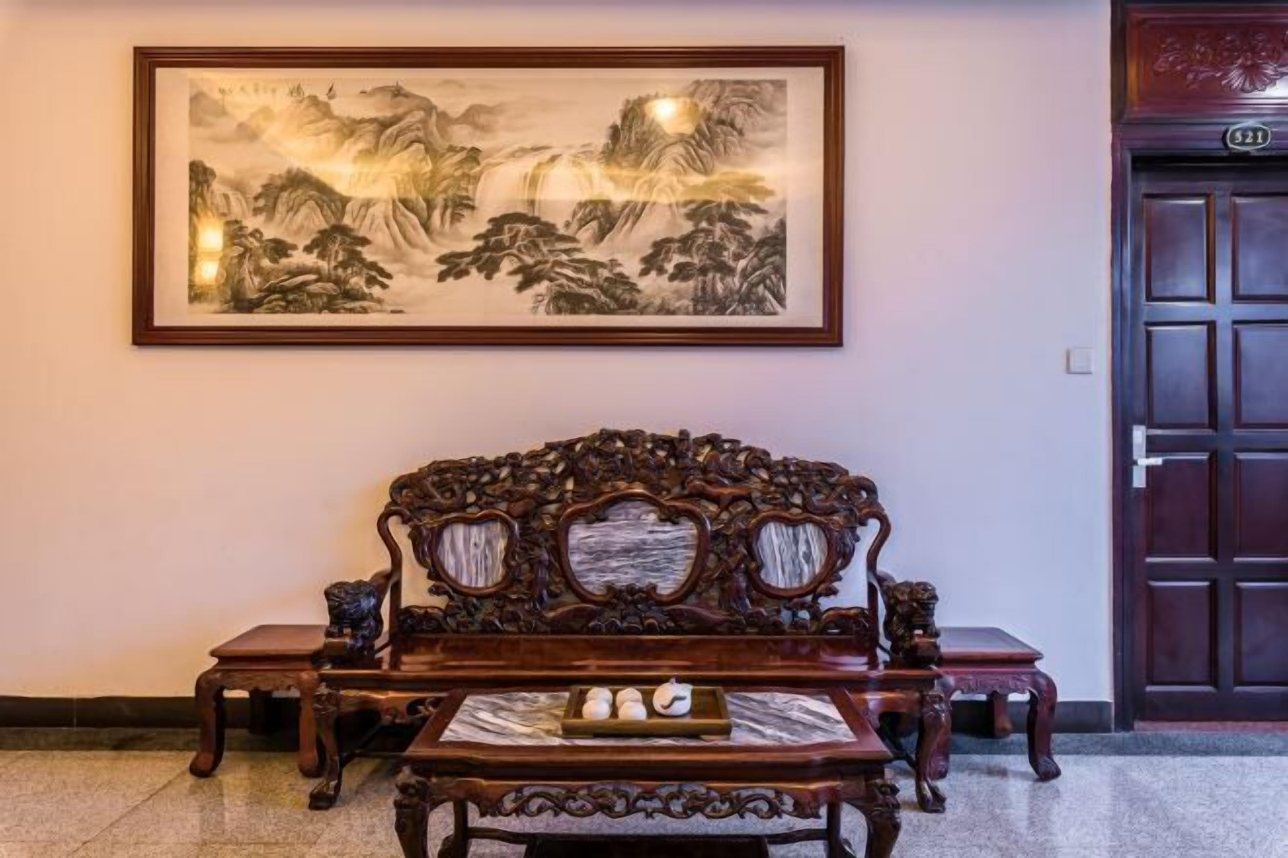 A traditional wooden sofa with intricate carvings, a framed landscape painting on the wall, and a wooden door with a number plate.
