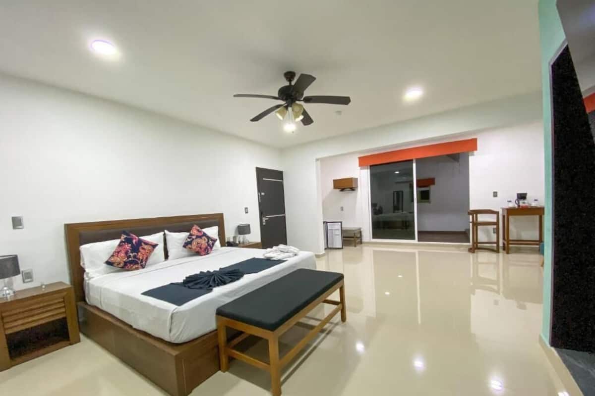 A modern hotel room with a large bed, bedside tables, a bench, a ceiling fan, and a sliding door leading to a balcony.