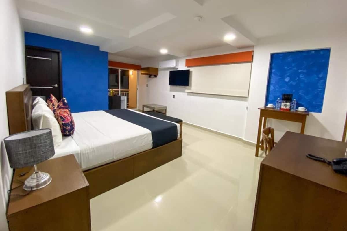A hotel room with a bed, bedside table, lamp, TV, and a small desk with a chair.