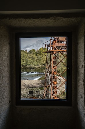 A window view of a metal structure by a river with trees and a cloudy sky.