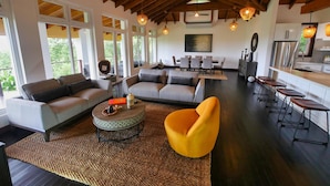 A spacious living area with a high ceiling, large windows, and a modern kitchen. - Relax at this hillside retreat that overlooks the lush forest and the Caribbean. (West End)