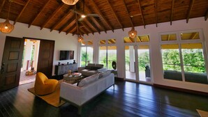 Amplia habitación con techo alto, vigas de madera, un sofá, una silla, una mesita y grandes ventanales con vistas a zonas verdes.