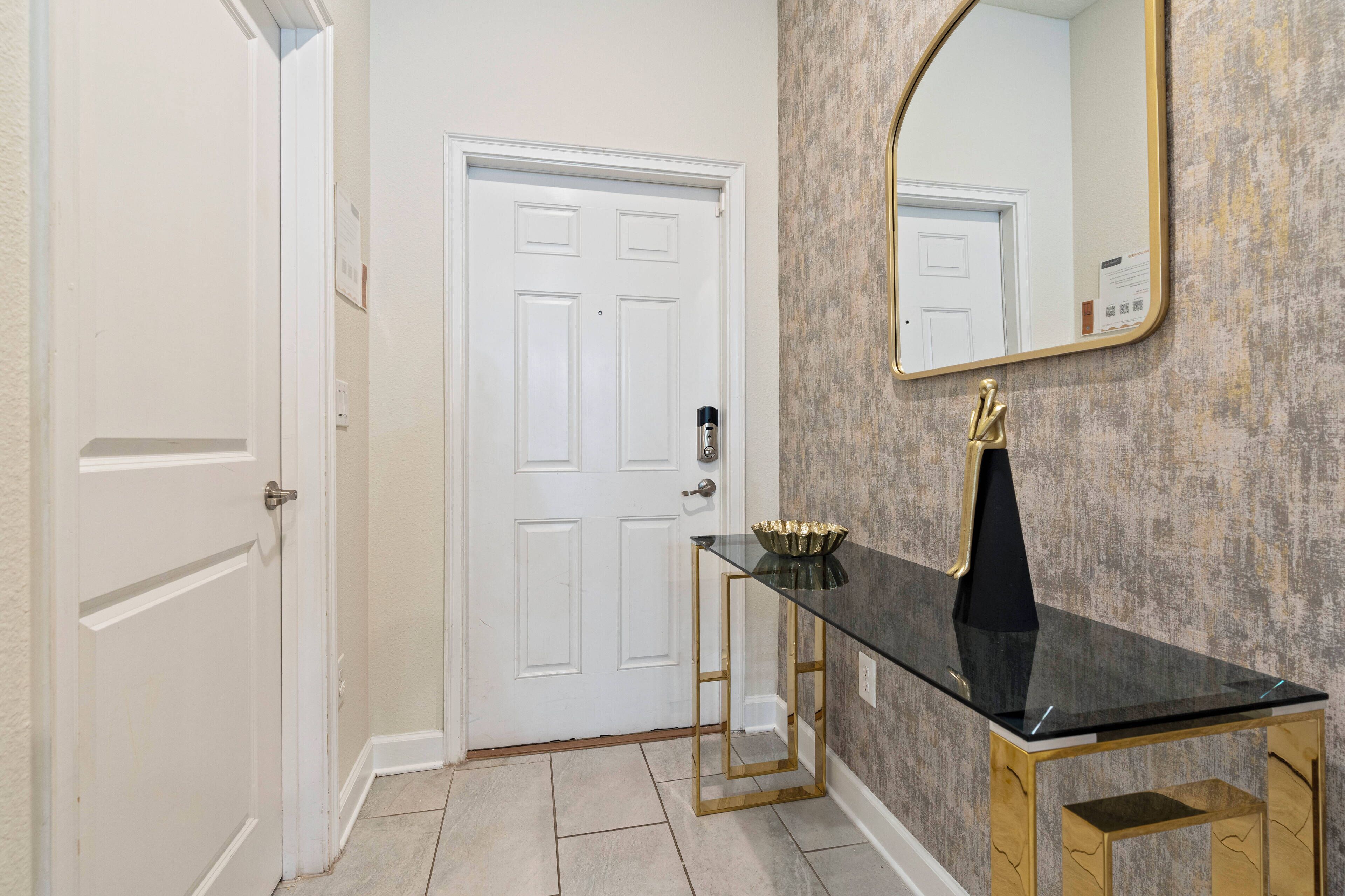 A hallway with a white door, a glass-top console table with gold legs, and a large mirror on the wall.