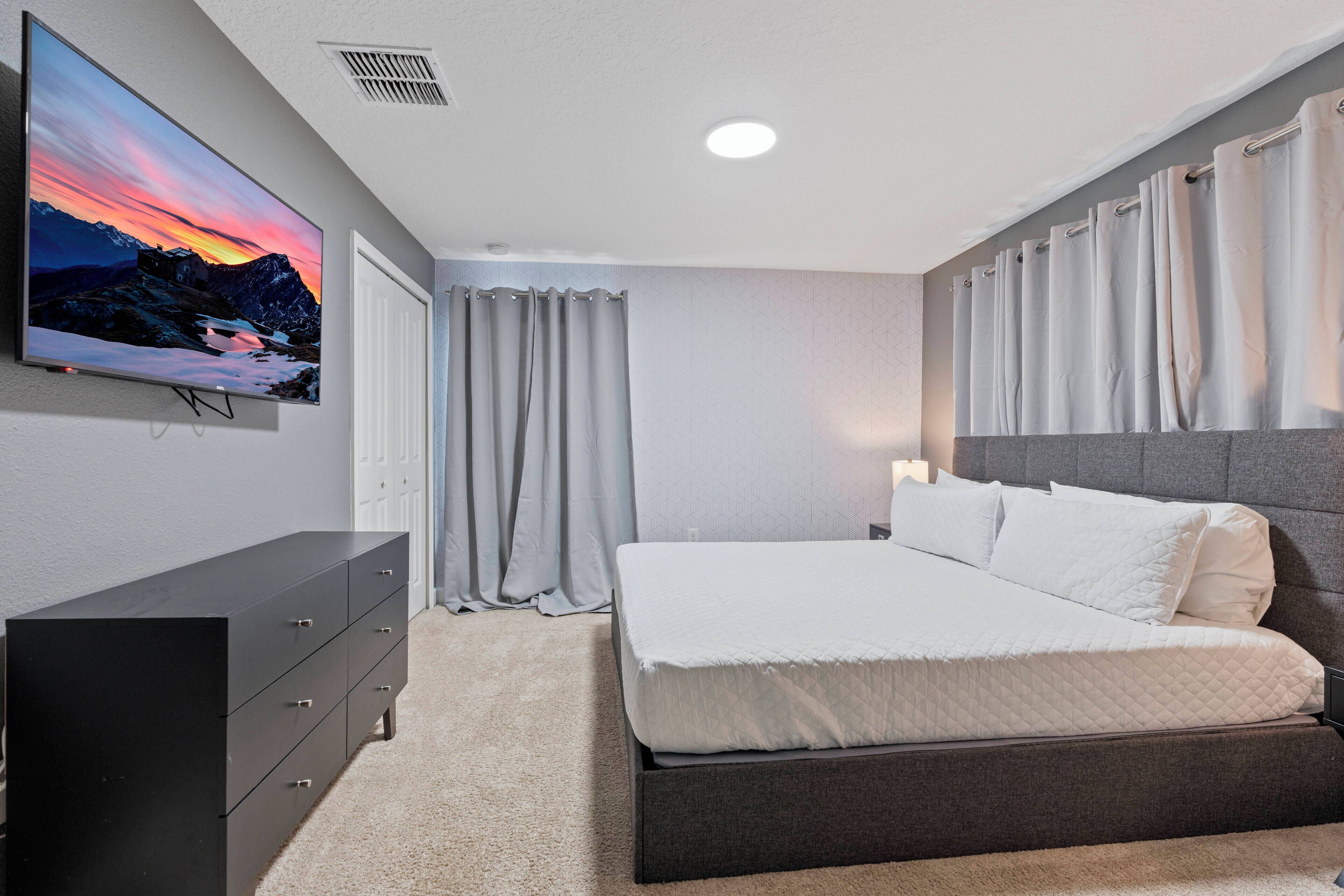 A modern bedroom with a large bed, a flat-screen TV mounted on the wall, and a dark dresser.