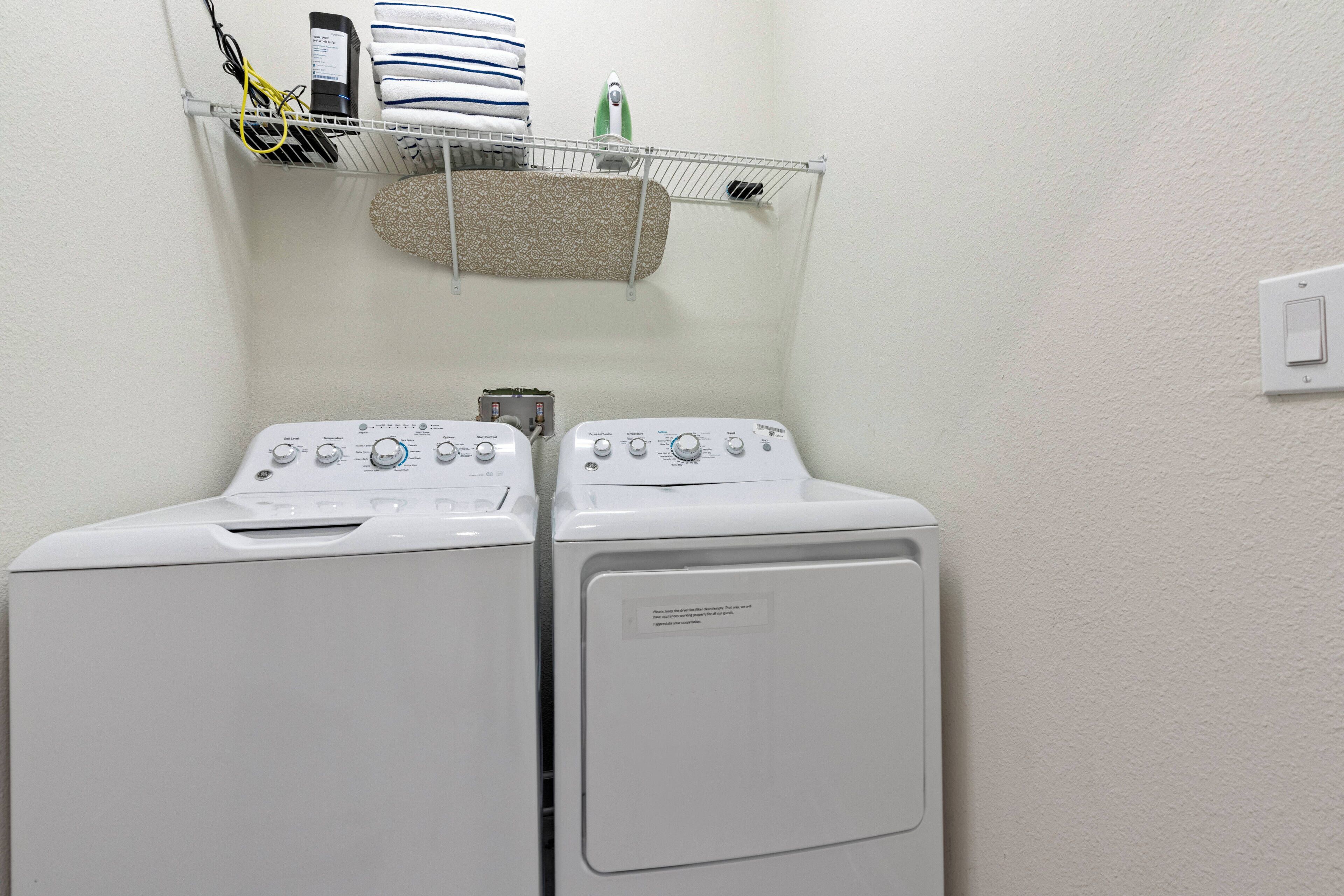 A laundry area with two stacked front-loading washing machines, a shelf with folded towels, and a hanging rack with laundry items.