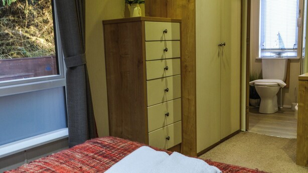 A bedroom with a bed, a wooden dresser, a window, and a bathroom with a toilet and sink. - Griffon Lodge - UK3469 (Felton, near Morpeth)