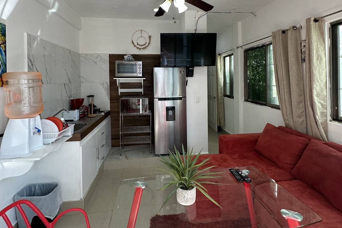 Un salon moderne avec une table basse en verre, des fauteuils rouges, un canapé rouge, un ventilateur de plafond, un téléviseur et un coin cuisine équipé d’un réfrigérateur, d’un four à micro-ondes et d’un distributeur d’eau.