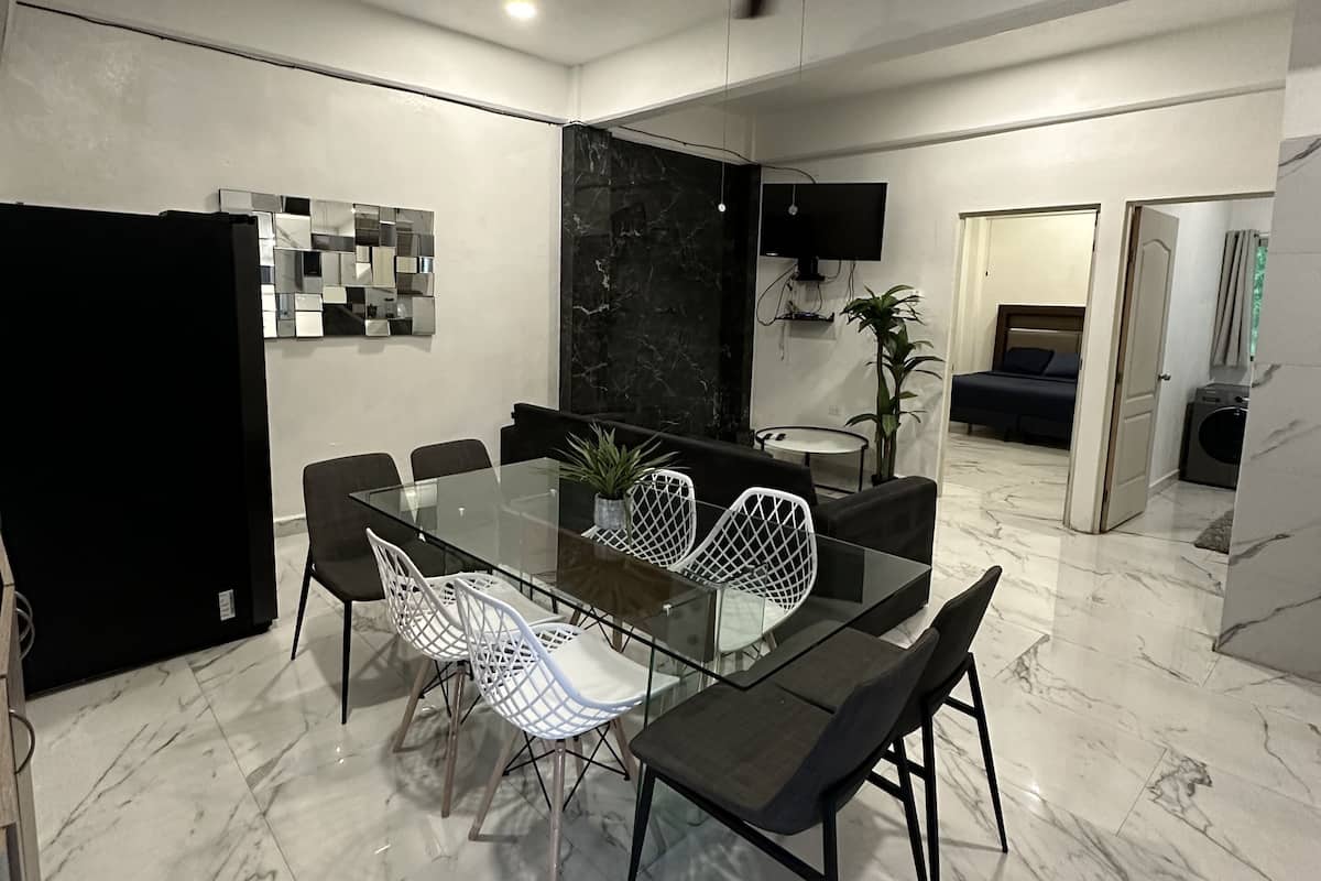 Un espace repas moderne avec une table en verre, des chaises noires et blanches, un ventilateur de plafond et un téléviseur fixé au mur.
