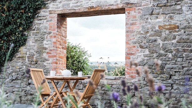 Un muro de piedra con una ventana, una mesa con sillas y vistas al campo.