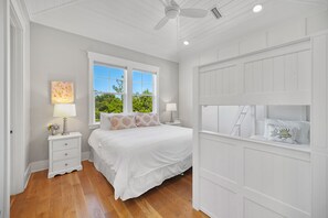 Quarto com cama, duas mesas de cabeceira, ventoinha no teto e janela com vista para árvores.