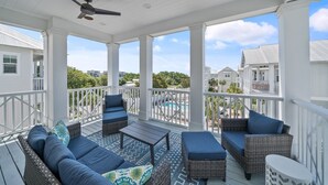 A covered outdoor deck with wicker furniture, a ceiling fan, and a view of a swimming pool and buildings. - Someday Came 4 Bedroom Home by RedAwning (Panama City Beach)