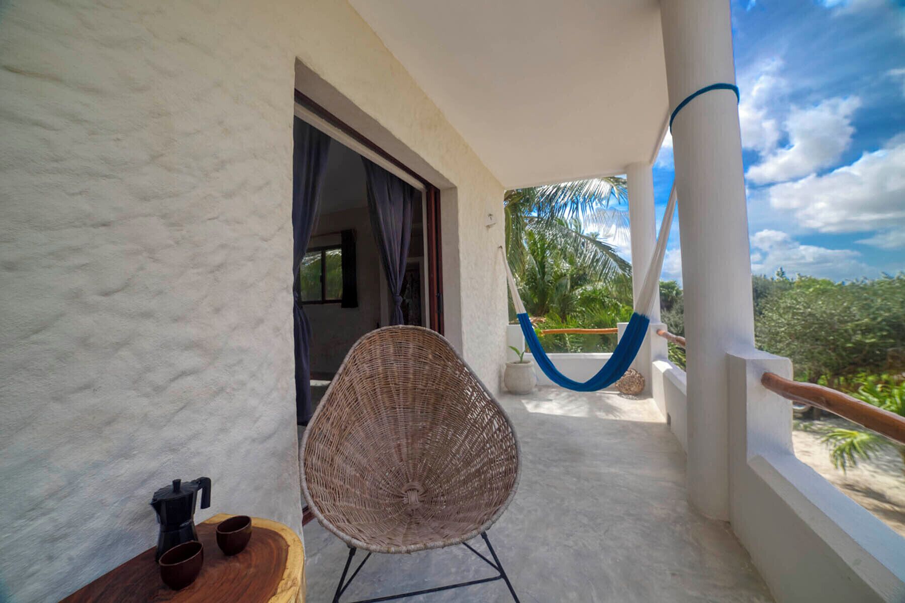 A balcony with a wicker chair, a small table with a coffee maker, and a view of palm trees and the sky.
