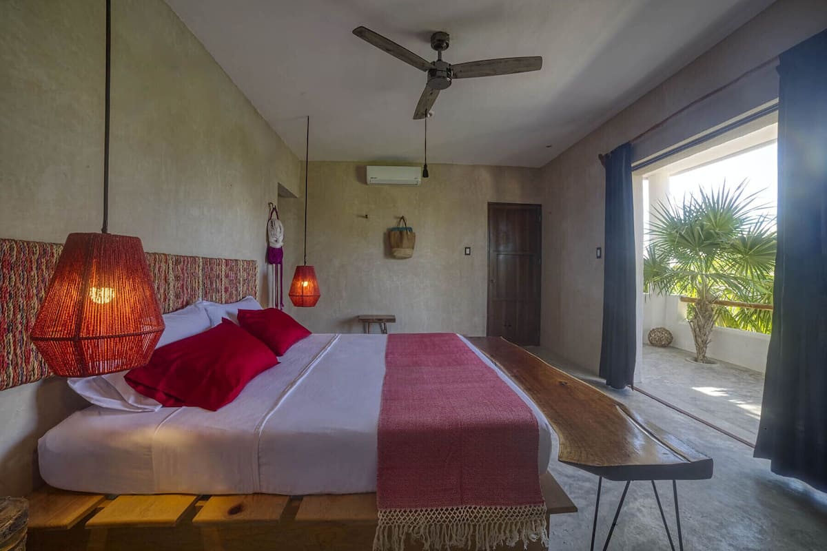 A bedroom with a bed, a ceiling fan, a hanging light fixture, and a view of the outdoors.