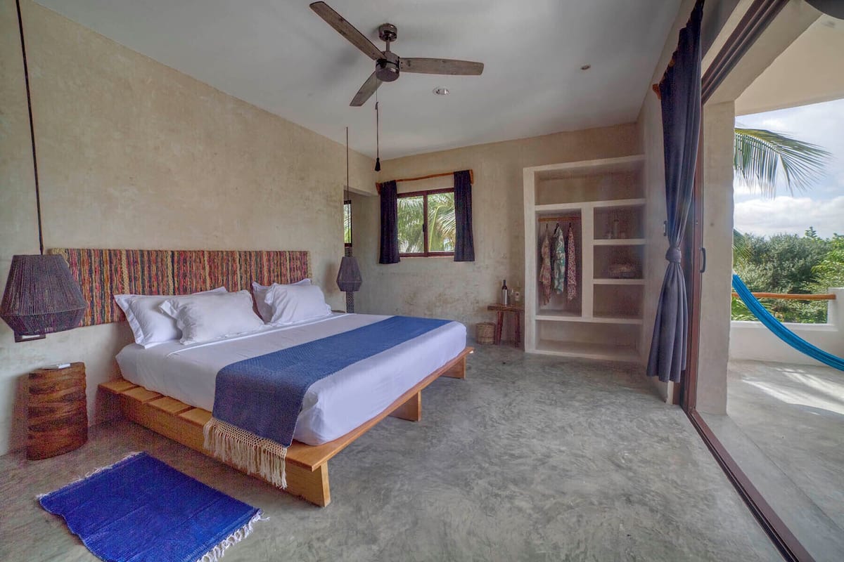 A bedroom with a bed, a ceiling fan, a window with curtains, a shelf with clothes, and a view of greenery outside.