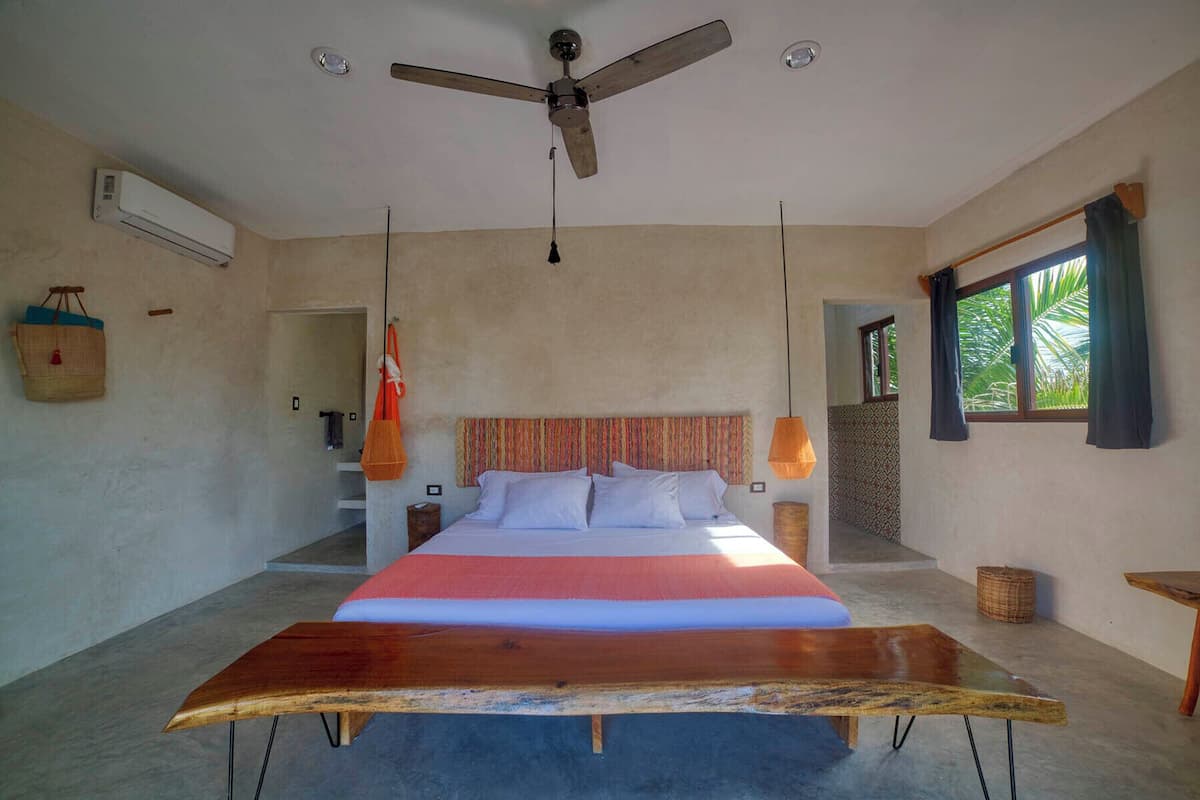 A bedroom with a large bed, a ceiling fan, an air conditioning unit, a window with curtains, and a view of palm trees.