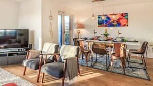 A modern living room with a TV, seating, and a dining area with a table set for four. - Marram Dune Beach House (Brean)