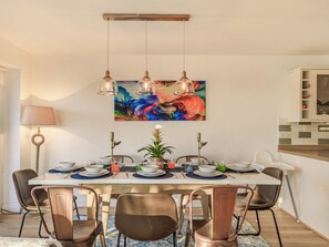 Uma área de refeições com uma mesa posta para seis pessoas, rodeada por seis cadeiras, um quadro na parede e uma área de cozinha à direita.