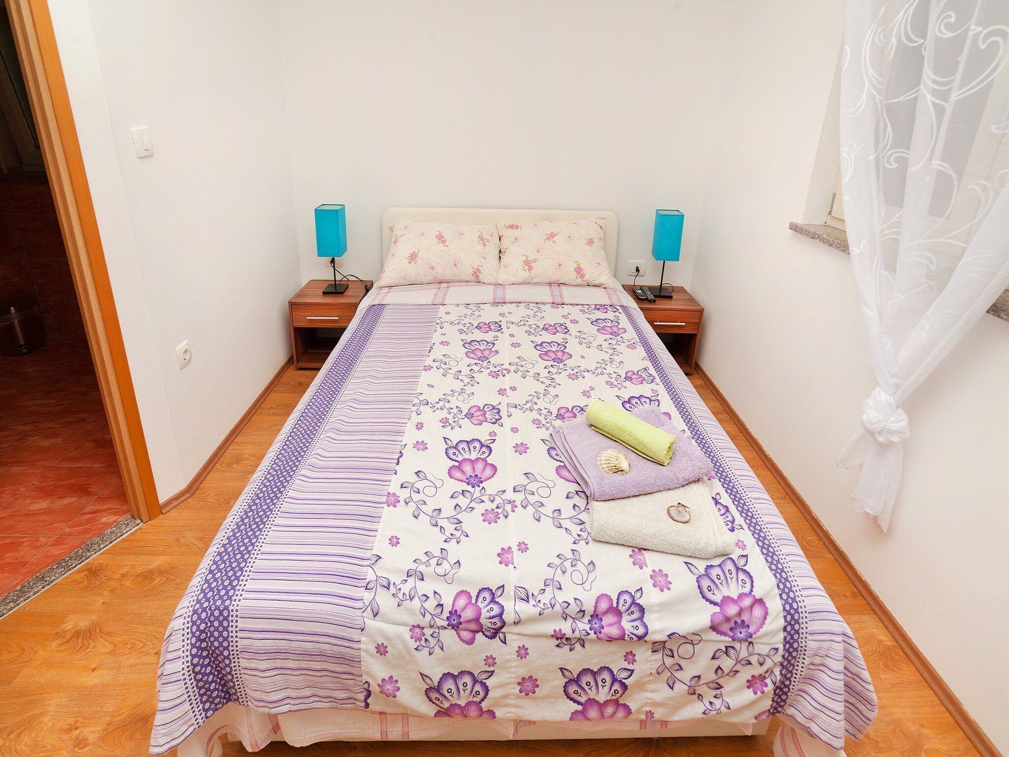 A neatly made bed with floral bedding, two bedside tables with lamps, and a window with sheer curtains.