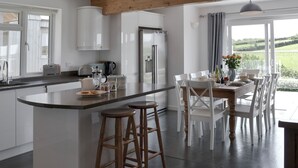 Eine moderne Küche mit weißen Schränken, einer zentralen Kochinsel inklusive Barhocker, einem Essbereich mit Tisch und Stühlen und Blick ins Grüne durch große Fenster.