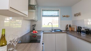 Una cocina compacta con armarios blancos, una ventana con cortina azul, un lavamanos y una encimera con una botella de vino y copas.