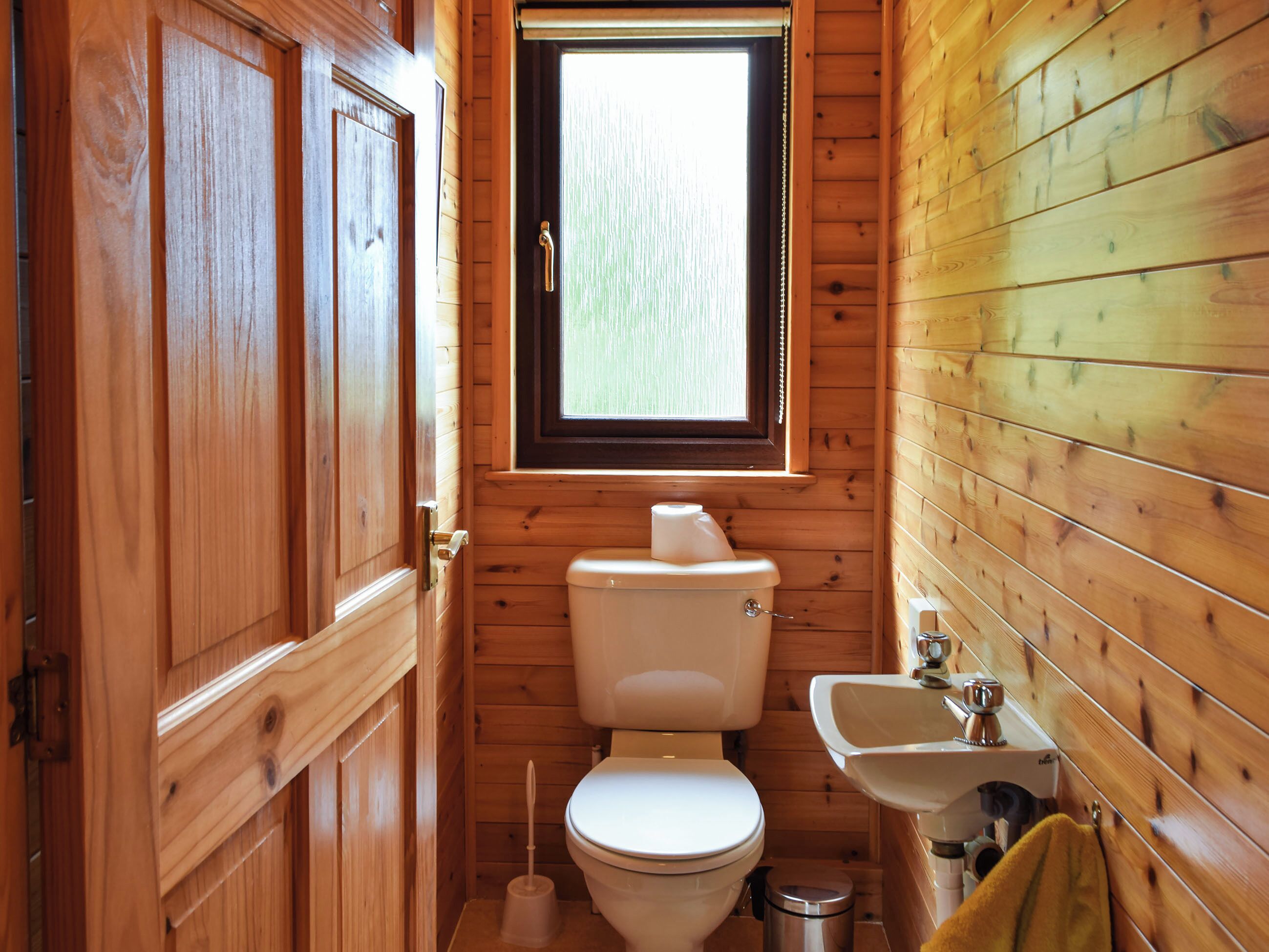 A compact wooden bathroom with a toilet, sink, and window.