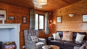 Un intérieur de chalet chaleureux avec une cheminée, un fauteuil à carreaux, un canapé en cuir et un tapis coloré.