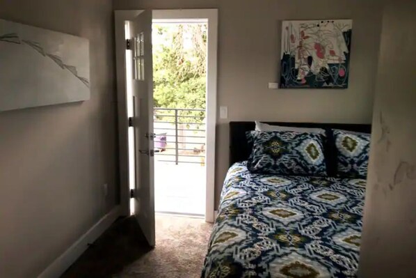 A bedroom with a patterned bedspread, a door leading to an outdoor area, and a wall-mounted abstract painting.