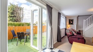 Une pièce avec un sol à carreaux, une table en bois, un canapé rouge et une vue sur une pelouse verdoyante et une clôture en bois, visibles à travers de grandes portes-fenêtres coulissantes.