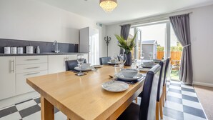 Un coin repas avec une table en bois dressée pour quatre personnes, une cuisine en arrière-plan et une vue sur le jardin à travers une porte ouverte.