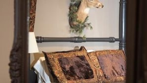 Una habitación con una cama, una lámpara y un cojín estampado, y un ciervo montado en la pared sobre la cama.